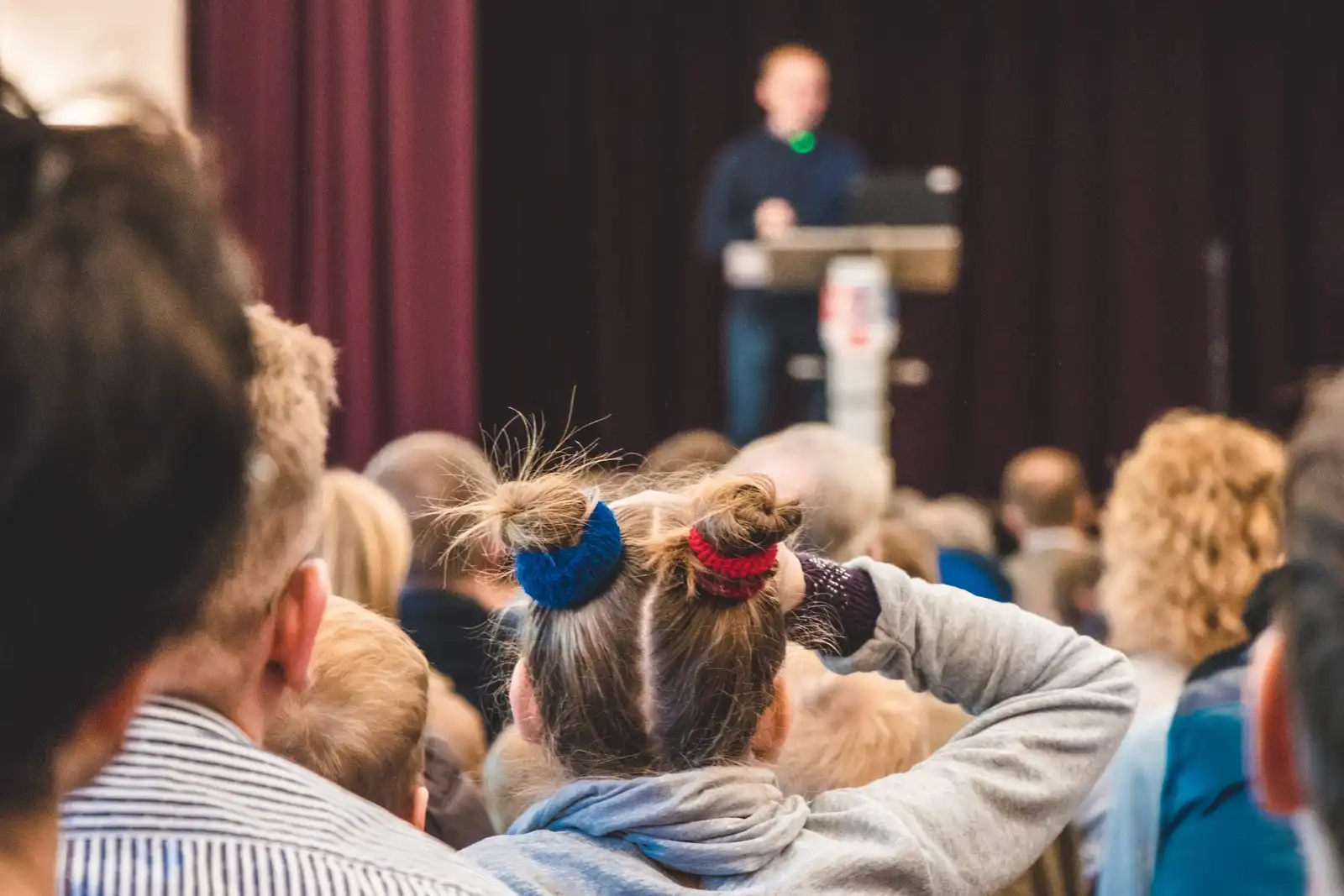 An audience seen from behind, listening to a speaker at a lectern