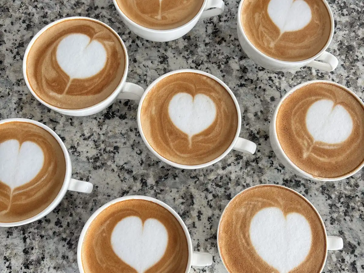 Eight cups of latte with heart-shaped foam on a speckled grey counter