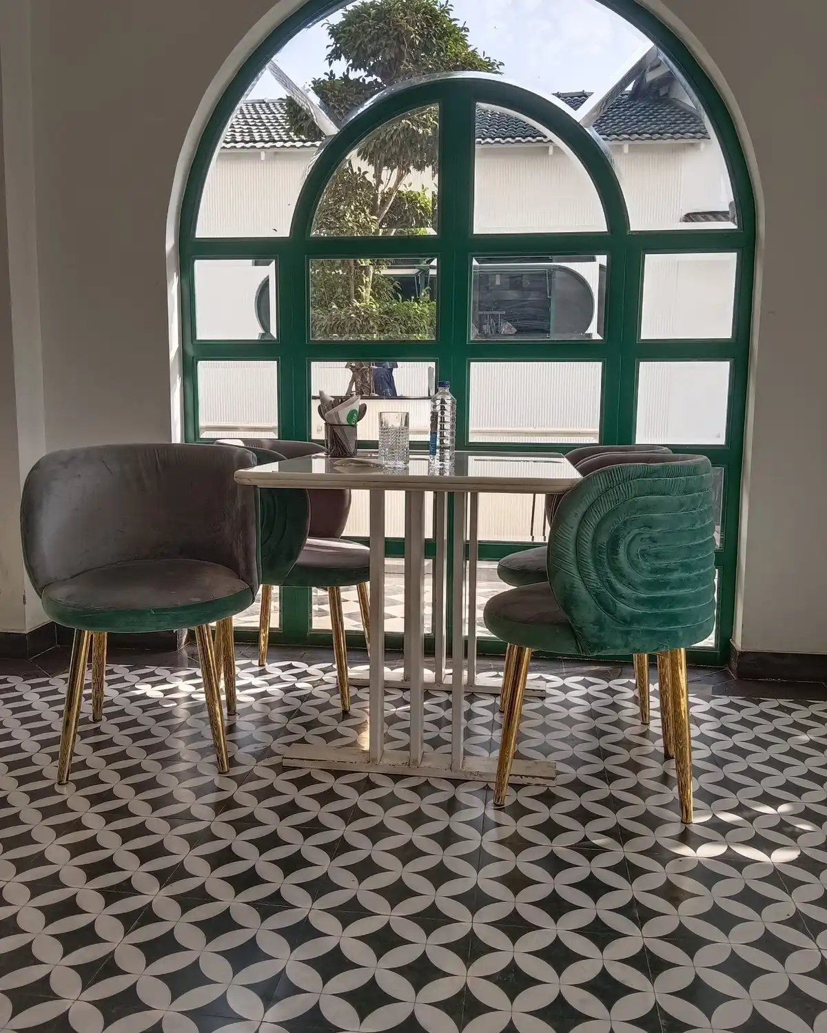 A café table with green velvet chairs in front of an arched window, on a patterned tile floor